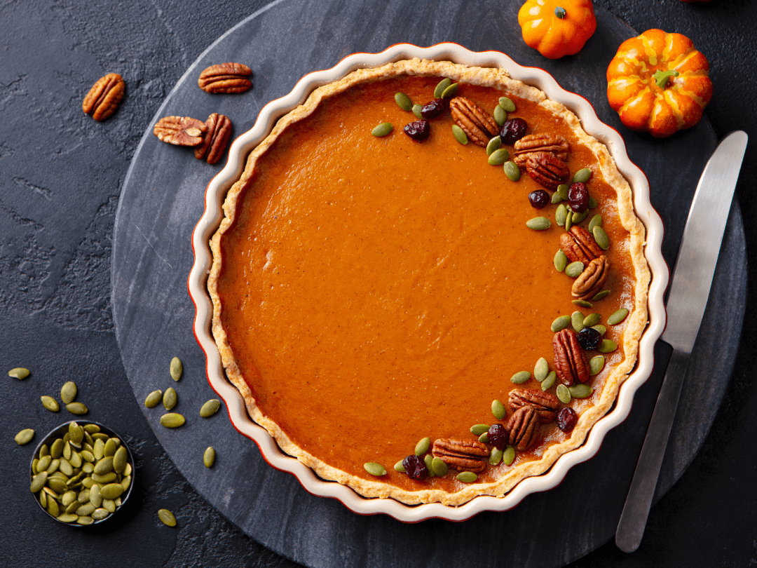 Pumpkin pie topped with pecans and pumpkin seeds in a white and red crust, on a dark serving tray.